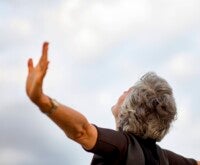 Mulher idosa de braços abertos olhando para o céu.